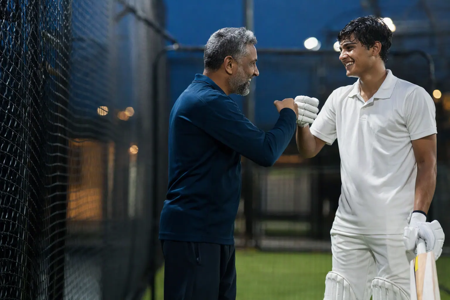 A cricket coach and a young batter bump fists after a good shot in the nets